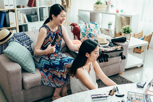 mujer peluquería amiga por plancha de pelo en el sofá en casa. dos chicas asiáticas riendo charlando mientras se maquillan se preparan por la mañana para viajar de verano a la playa. amistad feliz estilo de vida