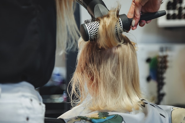 La mujer del peluquero hace el peinado bonito de moda para la linda niña rubia en la peluquería moderna, peluquería. Peluquero hace peinado para bebé en peluquería. Concepto de peinado y belleza