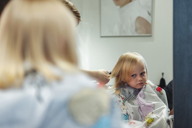 La mujer del peluquero hace el peinado bonito de moda para la linda niña rubia en la peluquería moderna, peluquería. Peluquero hace peinado para bebé en peluquería. Concepto de peinado y belleza