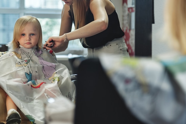 La mujer del peluquero hace el peinado bonito de moda para la linda niña rubia en la peluquería moderna, peluquería. Peluquero hace peinado para bebé en peluquería. Concepto de peinado y belleza