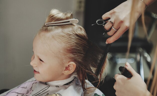 La mujer del peluquero hace el peinado bonito de moda para la linda niña rubia en la peluquería moderna, peluquería. Peluquero hace peinado para bebé en peluquería. Concepto de peinado y belleza