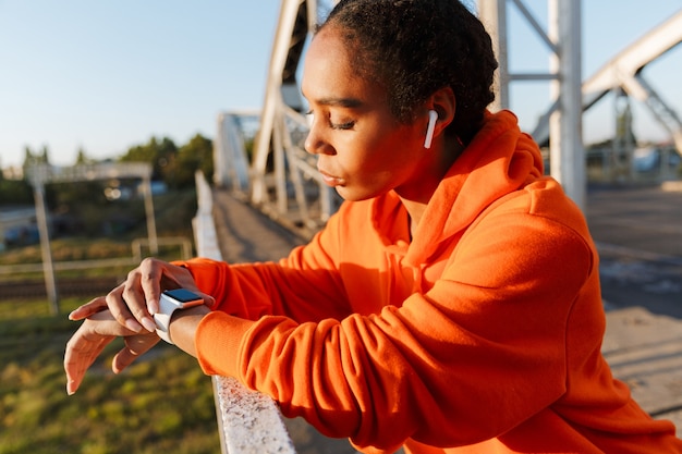 Mujer de pensamiento afroamericano en ropa deportiva con earpods y cronómetro mientras se apoya en la barandilla en el puente viejo