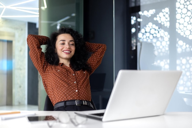 Mujer pensante feliz en el lugar de trabajo dentro de la oficina mujer de negocios con las manos detrás de la cabeza trabajo completado