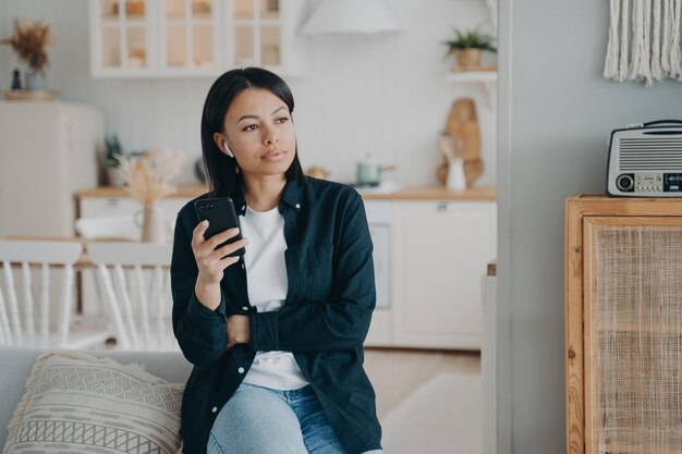 Mujer pensativa en auriculares con teléfono inteligente escuchando podcast de música o audiolibro en casa