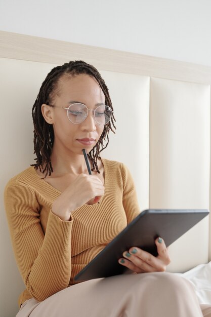 Mujer pensativa con gafas trabajando en un artículo para el periódico local que está revisando el texto en la tableta c ...