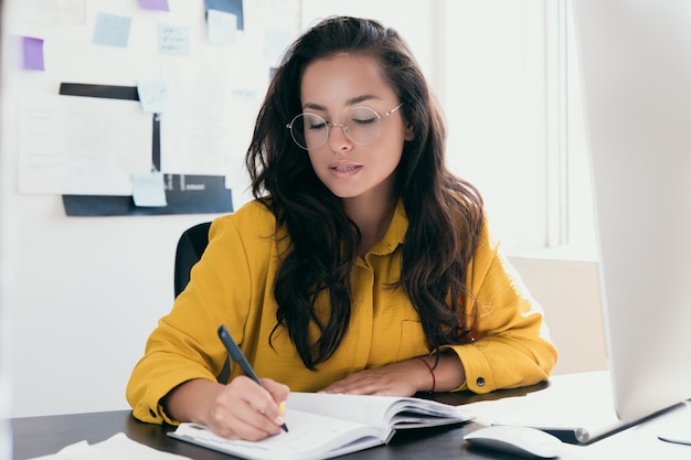 Mujer pensativa en el horario de trabajo de planificación de gafas escribiendo en el cuaderno mientras está sentado en el lugar de trabajo