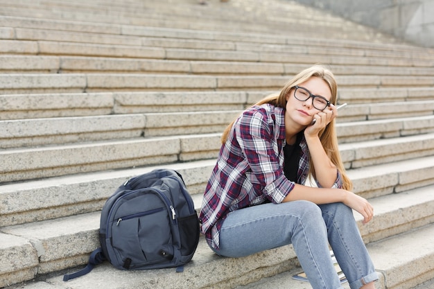 Mujer pensativa joven sentada al aire libre en las escaleras de la universidad con smartphone. Concepto de tecnología, educación y trabajo remoto, espacio de copia