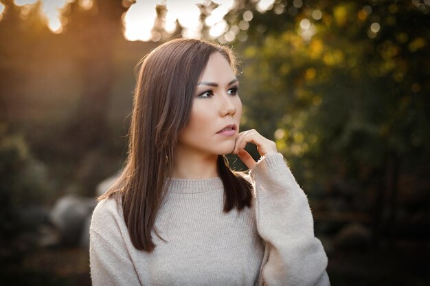 Mujer pensativa con la mano cerca de la cara en el parque al aire libre entre árboles al atardecer