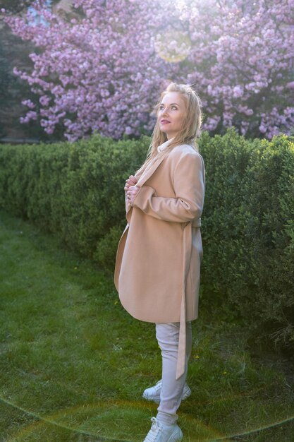 Mujer pensativa de mediana edad con abrigo beige y cabello rubio largo en el parque de primavera Retrato en toda longitud Marco vertical