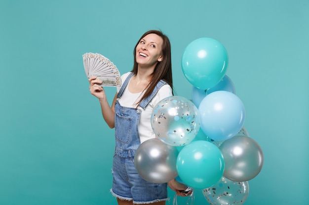 Una mujer pensativa mirando hacia arriba sostiene un abanico de dinero en billetes de dólar dinero en efectivo celebrando con coloridos globos de aire aislados en un fondo azul turquesa. Fiesta de cumpleaños, concepto de emociones de la gente.