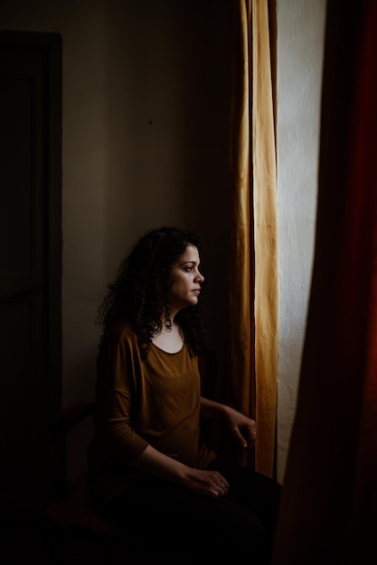 Mujer pensativa mirando por la ventana mientras está de pie en la oscuridad en casa