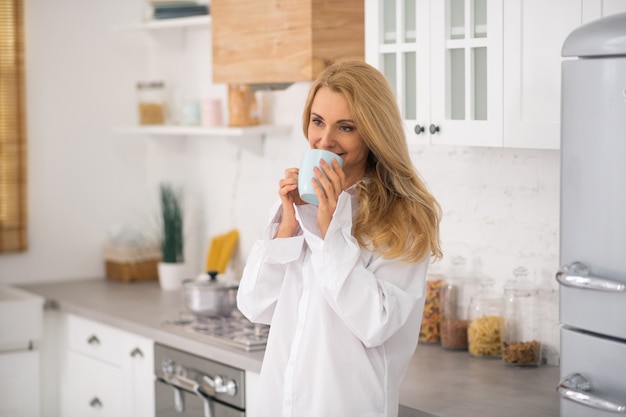 Mujer pensativa muy sonriente con taza cerca de la cara en casa en la cocina moderna blanca