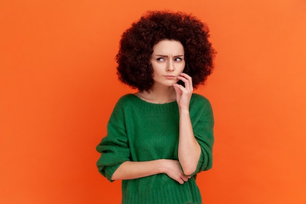 Mujer pensativa con peinado afro con suéter verde de estilo casual pensando, mirando hacia otro lado, con una expresión facial confundida y desconcertada. Disparo de estudio interior aislado sobre fondo naranja.