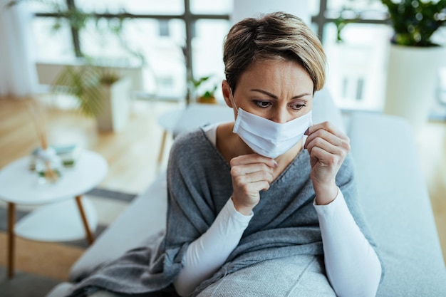 Mujer pensativa que se siente preocupada y sentada en casa con una máscara protectora en la cara durante la epidemia de virus