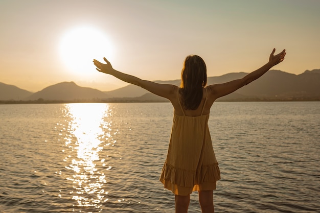 Mujer pensativa solitaria con los brazos abiertos en adoración del sol reflejándose en el agua de mar al atardecer o al amanecer