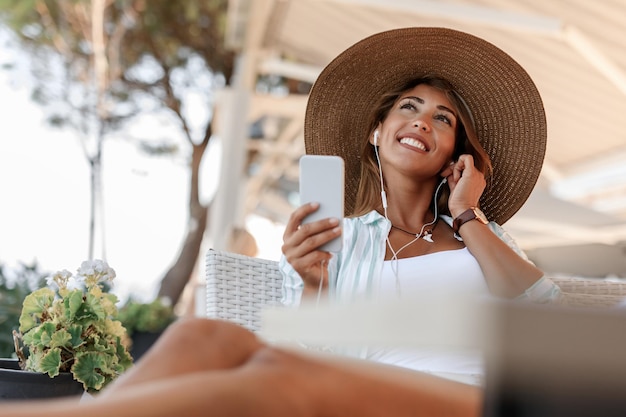 Mujer pensativa sonriente usando un teléfono inteligente y escuchando música mientras se relaja en un café