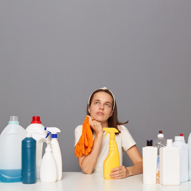 Una mujer pensativa usa una banda para la cabeza y una camiseta casual aislada sobre un fondo gris rodeada de muchos productos de limpieza mirando el espacio de la copia para publicidad