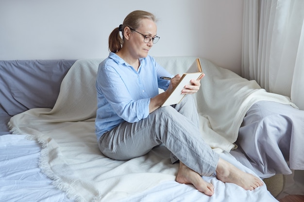 Mujer pensionista con gafas leyendo un libro, tomando notas con un lápiz mientras está sentado en el sofá en casa.