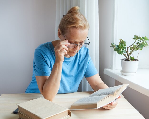 Mujer pensionista lee un libro junto a la ventana, ve mal con gafas.