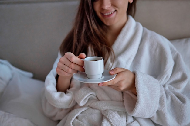 Mujer perezosa en bata de baño acostada en la cama y disfrutando de un buen desayuno con una aromática taza de café durante el descanso en la acogedora habitación de confort en la habitación del hotel