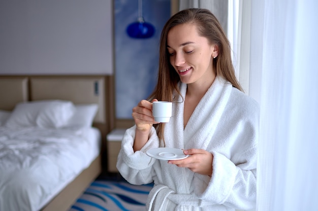 Mujer perezosa en bata de baño disfrutando de buenos días con aromática taza de café durante el descanso en la acogedora habitación de confort en la habitación del hotel