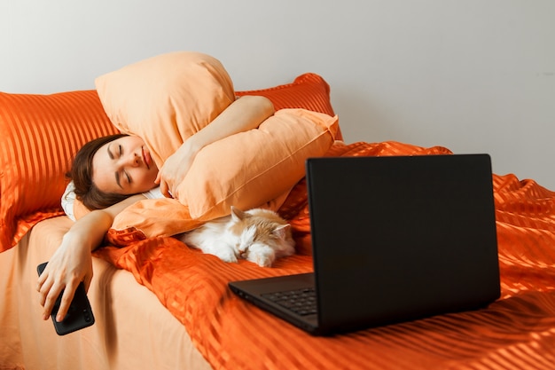 Una mujer perezosa duerme en una cama con una computadora portátil sobre sus rodillas y un gato dormido cerca.