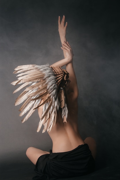 Mujer perfecta desnuda en el atuendo de indios americanos en el humo en una pared gris. Sombrero hecho de plumas. Misteriosa forma mística, un cuerpo sexy, una espalda hermosa