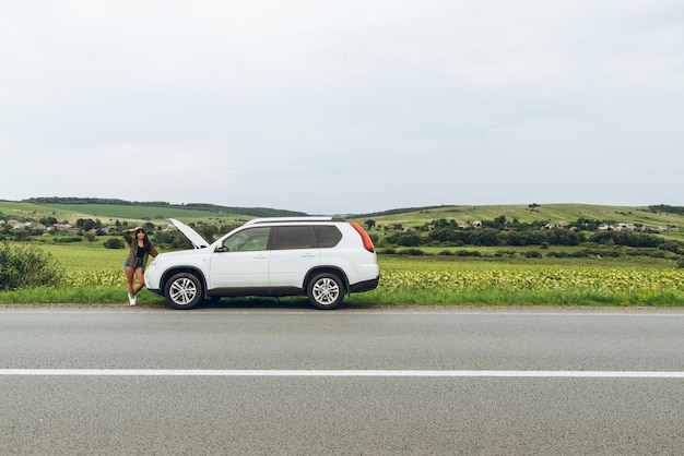 Mujer de pie al lado de la carretera cerca de un coche roto con el capó abierto. accidente en la carretera