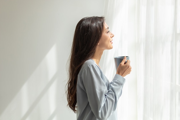 Mujer de pie beber café y abrir las cortinas de la ventana respirar aire fresco ejercicio de estiramiento en el dormitorio