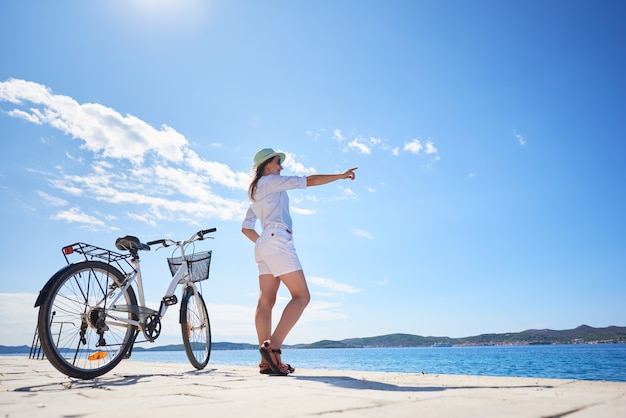 Mujer de pie en bicicleta en la acera pedregosa bajo un cielo azul claro apuntando a la orilla opuesta sobre fondo de agua clara y brillante