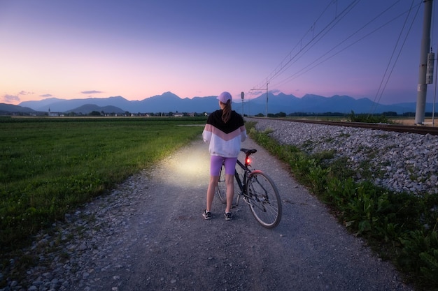 Foto la mujer está de pie con una bicicleta de montaña en un camino de ripio por la noche en verano paisaje colorido con una bicicleta deportiva con campo de luz camino de tierra hierba verde cielo púrpura al atardecer deporte y viajes