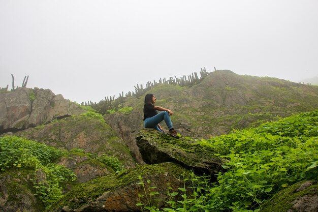 Mujer de pie encima de una roca alrededor de plantas verdes fondo montañoso y cielo nublado