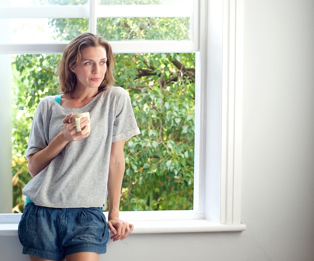 Mujer de pie junto a la ventana en casa con una taza de café