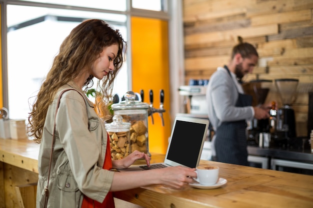 Mujer de pie en el mostrador y usando laptop mientras tomando un café