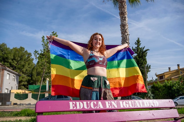 Una mujer de pie en la parte superior de un banco rosa sosteniendo una bandera arco iris