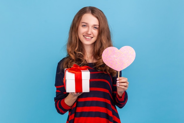 Mujer de pie sosteniendo una caja de regalo y mostrando un corazón rosa en un palo mirando a la cámara con amor