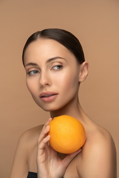 Mujer con piel facial perfecta, cabello peinado, sosteniendo naranja