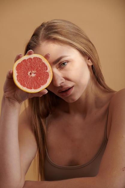 Foto mujer de piel limpia sonriente feliz sosteniendo pomelo rojo cerca de la cara