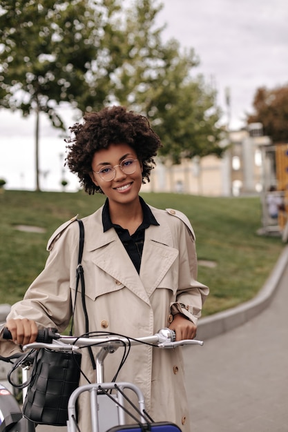 Mujer de piel oscura de ojos marrones en gabardina beige sonríe sinceramente y posa con bicicleta al aire libre