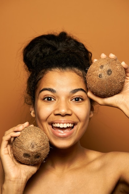 Foto mujer con piel perfecta y cabello en un moño sosteniendo dos cocos cerca de su rostro, posando aislado en naranja