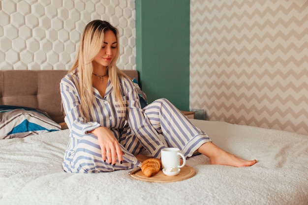 Una mujer en pijama bebe café con un croissant en la cama