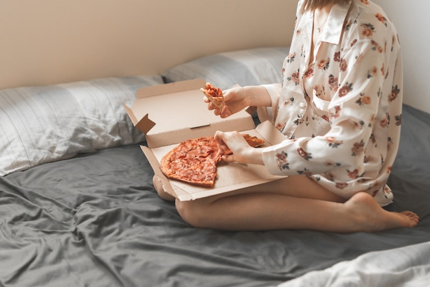 Mujer en pijama sentada en la cama, sostiene una caja de pizza sobre sus rodillas