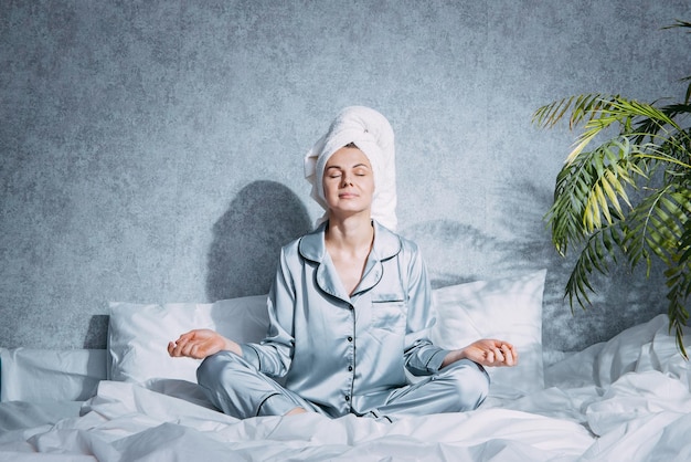 Una mujer en pijama y una toalla en la cabeza se sienta en una cama bajo el sol de la mañana en posición de loto y medita