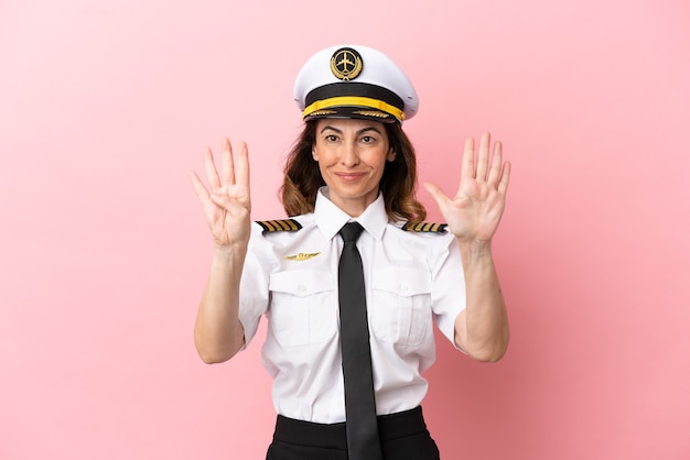 Mujer piloto de mediana edad de avión aislada sobre fondo rosa contando nueve con los dedos