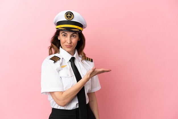 Mujer piloto de mediana edad de avión aislada sobre fondo rosa presentando una idea mientras mira sonriendo hacia