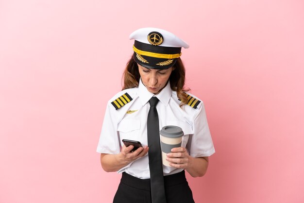 Mujer piloto de mediana edad de avión aislada sobre fondo rosa sosteniendo café para llevar y un móvil