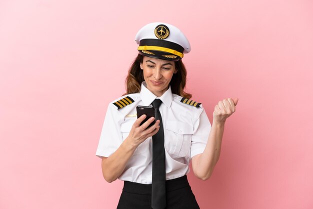 Mujer piloto de mediana edad de avión aislada sobre fondo rosa con teléfono en posición de victoria
