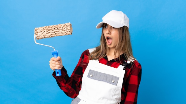 Mujer pintora sobre pared azul haciendo gesto de sorpresa mientras mira a un lado