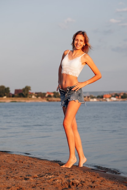 Una mujer en la playa una hermosa joven rubia sonriente con un top blanco de encaje y pantalones cortos de mezclilla desabrochados ...