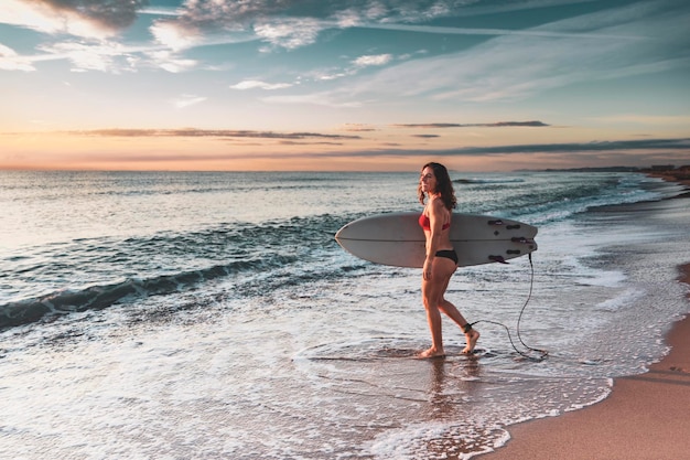 Mujer en la playa con tabla de surf entra en la playa en bikini al amanecer